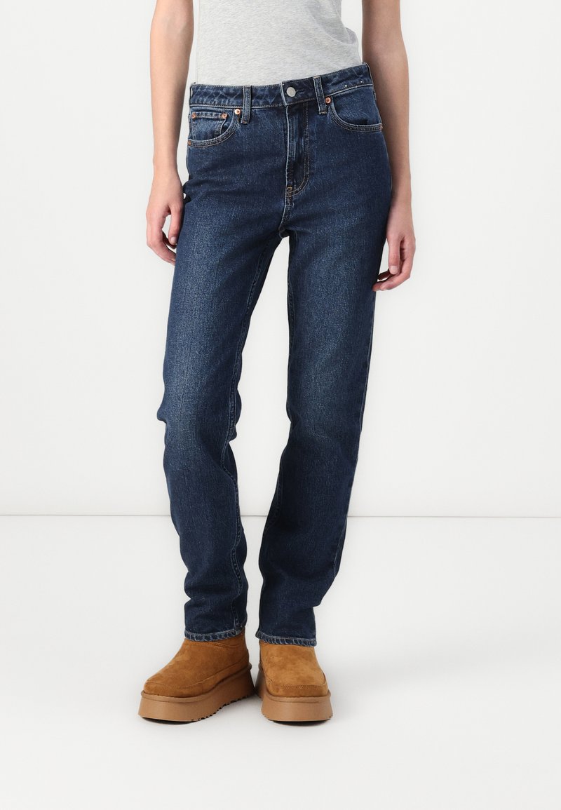 High-waisted, straight-leg jeans in dark blue denim, featuring subtle fading, five pockets, and copper-tone hardware. Paired with tan suede boots.
