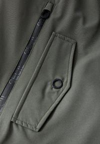 Tessuto della giacca verde oliva con una texture liscia, dotata di una tasca con zip e di una tasca a pattina cucita con un dettaglio hardware circolare nero.