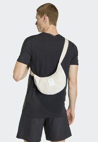 Bolso bandolera en beige claro, hecho de tela suave. Cuenta con una forma redonda, correa ajustable y una etiqueta con el logo en el frente. Se lleva en la espalda.