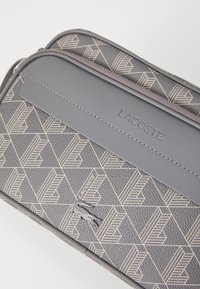 Pochette grigia con texture e motivo geometrico, caratterizzata da un pannello grigio liscio e logo "Lacoste" in rilievo, chiusa da una zip.