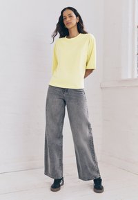 Gelbes Kurzarm-T-Shirt mit lockerem Schnitt, kombiniert mit hoch taillierten grauen Weitebein-Jeans und schwarzen Sneakern mit weißen Streifen.