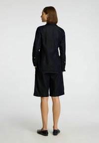 Outfit in denim scuro caratterizzato da una camicia a maniche lunghe con polsini a bottone e pantaloni corti fino al ginocchio, entrambi con una finitura testurizzata. Scarpe basse nere.