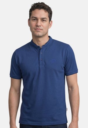 Homme aux cheveux courts et bouclés portant un t-shirt Henley bleu à manches courtes, debout devant un fond clair uni.