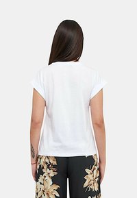 Maglietta bianca in cotone con maniche arrotolate, caratterizzata da un design semplice sul retro. Indossata con pantaloni scuri a fantasia floreale.