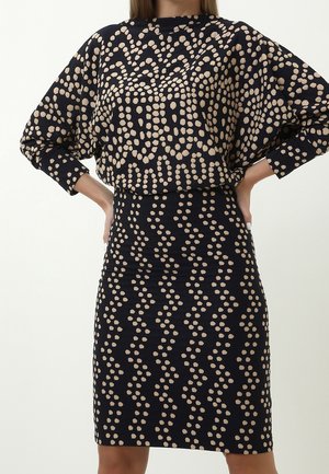 Kobieta ubrana w dopasowaną czarną spódnicę i luźną bluzkę z beżowymi abstrakcyjnymi kropkami, ręce opierające się na biodrach na jednolitym tle.