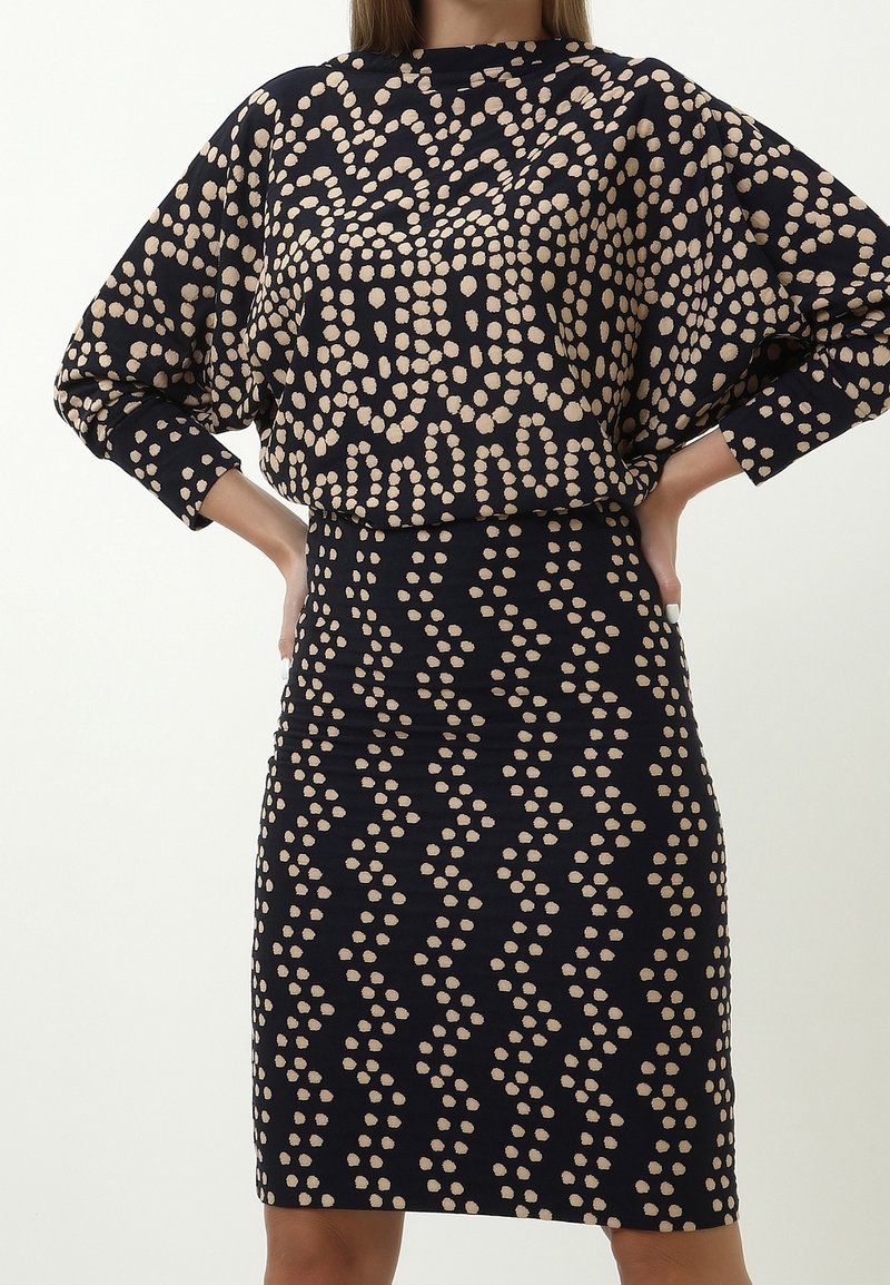 Femme portant une jupe noire ajustée et un haut ample avec des motifs abstraits en points beiges, mains posées sur les hanches sur un fond uni.