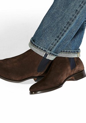 Botines de ante marrón con paneles laterales elásticos y punta afilada, combinados con jeans de denim azul arremangados.