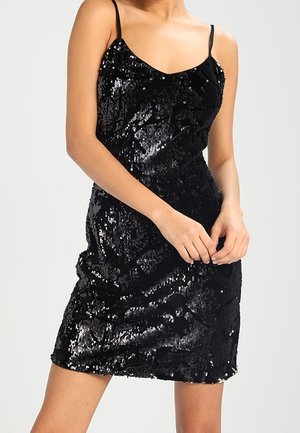 Femme portant une mini-robe noire sans manches couverte de sequins avec de fines bretelles et une silhouette ajustée.