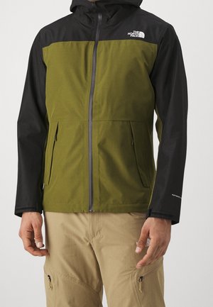Homme portant une veste à capuche North Face verte et noire avec un pantalon beige, debout, les bras détendus le long du corps.