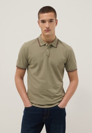 Giovane uomo con capelli corti che indossa una polo verde oliva con colletto a righe e jeans blu scuro, mani in tasca, in piedi su sfondo bianco.
