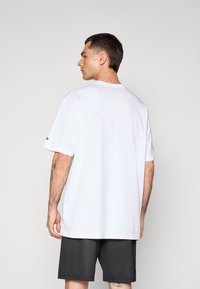 Camiseta blanca de manga corta con cuello redondo, fabricada en un tejido suave y con un corte holgado. Pantalones cortos de malla negros visibles en la parte inferior. Sin patrones o acentos distintos.