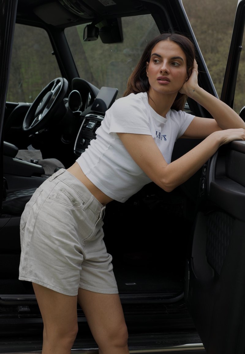 Weißes, kurzes T-Shirt mit blauer Schrift, kombiniert mit hellbeigen Shorts, lehnt an einer Fahrzeugtür. Das Innere ist sichtbar mit Armaturenbrett und Lenkrad.