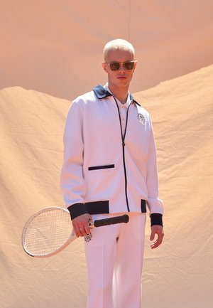 Witte zip-up jas met een marineblauwe kraag en zakaccenten, gedragen over een poloshirt; gecombineerd met op maat gemaakte witte broek. Houdt een tennisracket vast.