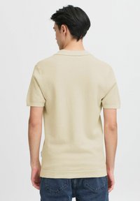 Beige rövid ujjú pólóing texturált anyagból, klasszikus gallérral és egyenes szabással, sötétkék farmernadrággal párosítva.