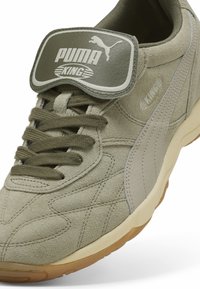 Sneaker Puma King en suède vert olive avec lacets épais, languette rembourrée affichant le logo Puma, et semelle en caoutchouc beige.