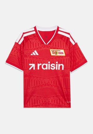 Maillot de football rouge avec des bandes blanches Adidas sur les épaules, logo de l'équipe 1. FC Union Berlin et texte du sponsor "raisin" sur le devant.