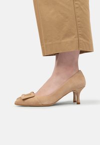 Pantof bej din suede cu toc înalt, cu vârful rotund și detaliu cu fundiță în față. Toc subțire, de înălțime medie. Textură simplă, netedă.