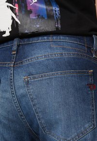 Tummansininen denimfarkku, jossa on sileä pinta, oranssit ompeleet ja punainen logo oikeassa taskussa. Klassinen viiden taskun malli.