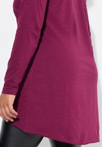 Pitkähihainen, löysä yläosa heather-magenta-kankaasta, jossa on kaareva helma, yhdistettynä tyylikkäisiin mustiin legginsseihin. Sujuva tekstuuri.
