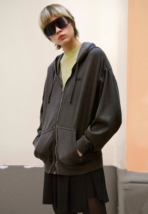 Dunkelgrau Zip-Hoodie mit einer Fronttasche und einer Kordelzugkapuze, kombiniert mit einem gerippten gelben Oberteil und einem schwarzen Plisseerock.