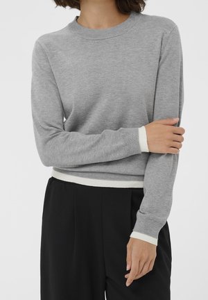Personne portant un pull gris clair à manches longues avec des poignets et un ourlet blancs, associé à un pantalon noir taille haute, bras croisés devant.