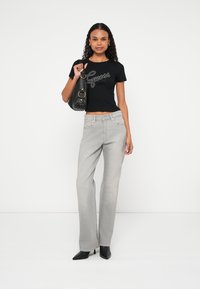 Czarny krótki T-shirt z logo "Guess", w połączeniu z jasnoszarymi, rozszerzanymi dżinsami. Modelka trzyma czarną torebkę i nosi czarne szpilki z ostrym noskiem.