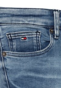 Jeans de mezclilla en un color azul desgastado, con un pequeño bolsillo frontal que presenta detalles en etiqueta rojo, blanco y azul, y con herrajes de botones plateados.