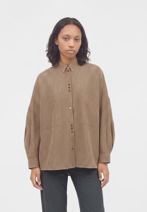 Femme aux cheveux mi-longs foncés portant une chemise ample à rayures marron boutonnée et un pantalon sombre, debout devant un fond clair uni.