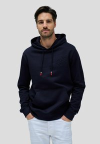 Felpa con cappuccio blu navy, dotata di tasca a marsupio, tessuto strutturato e logo in rilievo sul petto. Presenta puntali del cordoncino in rosso e bianco a contrasto.