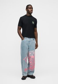 Άνδρας που φοράει μαύρο T-shirt με μικρό πράσινο γραφικό και ανοιχτό μπλε τζιν με μεγάλο ροζ γραφικό σχέδιο στο δεξί πόδι, στέκεται μπροστά σε λευκό φόντο.