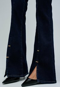Jeans de tiro alto en azul oscuro con detalles de costura coloridos y cortes laterales, combinados con unos tacones altos negros con punta.