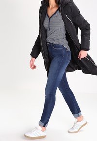 Svart vadderad parkas med huva, marinblå och vit randig henley tröja, blå skinny jeans och vita sneakers med gröna detaljer.