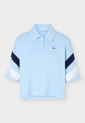 Tricou polo bleu deschis, scurtat, cu dungi diagonale bleumarin și albe pe mâneci și un mic logo cu un crocodil verde pe piept.