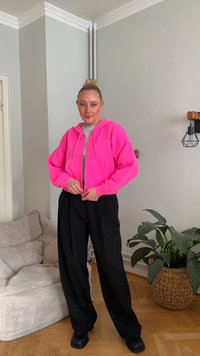 Felroze korte hoodie met trekkoord, grijs T-shirt eronder, gecombineerd met losse zwarte broek en chunky zwarte schoenen, binnenomgeving.