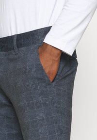 Gray textured pants with a check pattern, featuring a button closure and side pockets, paired with a white long-sleeve shirt.