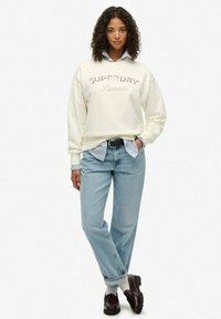 Sudadera crema con letras "SUPERDRY", combinada con una camisa de rayas azules, pantalones vaqueros de mezclilla azul claro y zapatos marrones.