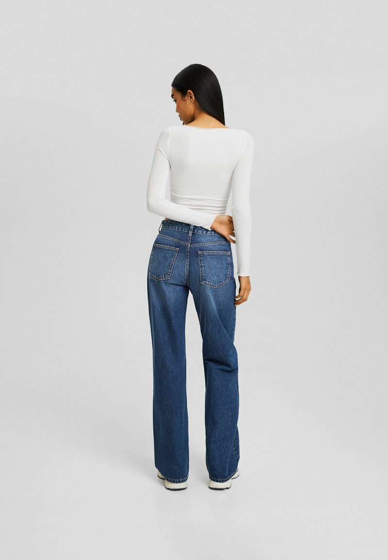 Leichtes, langärmliges, weißes Oberteil kombiniert mit locker sitzenden, dunkelblauen Denim-Jeans, die zwei Gesäßtaschen und eine gestickte Details aufweisen.