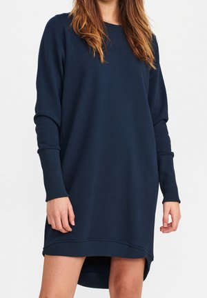 Kobieta nosząca granatową, długorękawową sukienkę typu sweatshirt z prążkowanymi mankietami i asymetrycznym dołem, stojąca na białym tle.