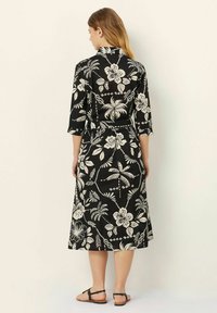 Abito floreale nero con maniche a 3/4, caratterizzato da una vita con fiocco e un design lungo e fluido con modelli tropicali bianchi a contrasto.