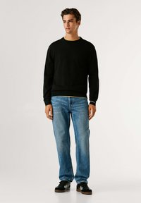 Črna crewneck kapuca z ribanim zapestjem, oblečena čez rumeno majico, v kombinaciji s svetlo modrimi kavbojkami in črnimi supergami. Gladka tekstura.