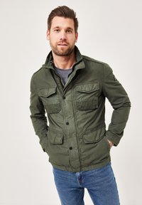 Olive green jacket made of cotton, featuring a zip and button closure, four front pockets, and a stand-up collar with stitching details.