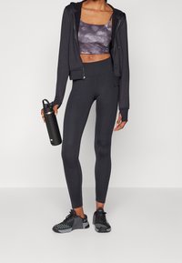 Sweatshirt noir zippé, crop top violet à motifs, leggings noirs taille haute et chaussures de sport noires. Tenant une bouteille d'eau noire.