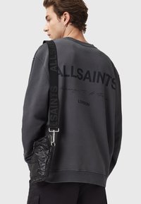 Grå sweatshirt med runde halsudskæring, prydet med stor sort tekst og et skriftskrift nedenfor. Sort crossover taske med en skinnende finish og justerbar rem.