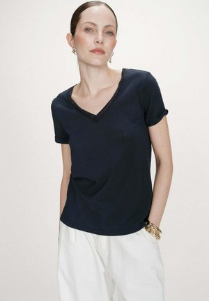 Femme portant un haut bleu marine à col en V et manches courtes avec revers, un pantalon blanc, accessoirisée de boucles d'oreilles anneaux en or et de plusieurs bracelets en or.