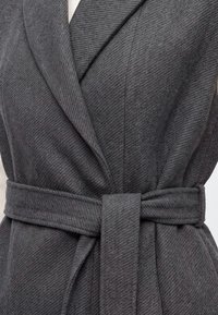 Gros plan d’un manteau en tissu chevron gris avec un large revers et une ceinture assortie nouée à la taille.