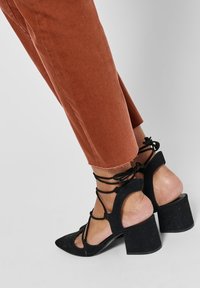 Chaussures à talons hauts noires à bout pointu, avec des brides lacées et un talon épais, associées à un pantalon court couleur rouille.