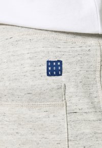 Sudadera gris claro hecha de material texturizado, con una etiqueta azul que dice "DNM WORKRS" y puños acanalados en las mangas y el dobladillo.