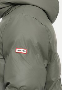Green puffer jacket with a smooth, shiny finish. Features a red and white "HUNTER" logo patch on the left sleeve.