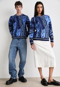Pulls en tricot bleu avec des motifs abstraits, associés à un jean bleu clair et une jupe blanche, le tout avec des chaussures à plateforme noires.