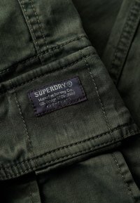 Primer plano de tela verde oscura con costuras cosidas y una etiqueta azul marino que dice "SUPERDRY® Manufacturing Co. GB Design 0106-3562" cosida al material.